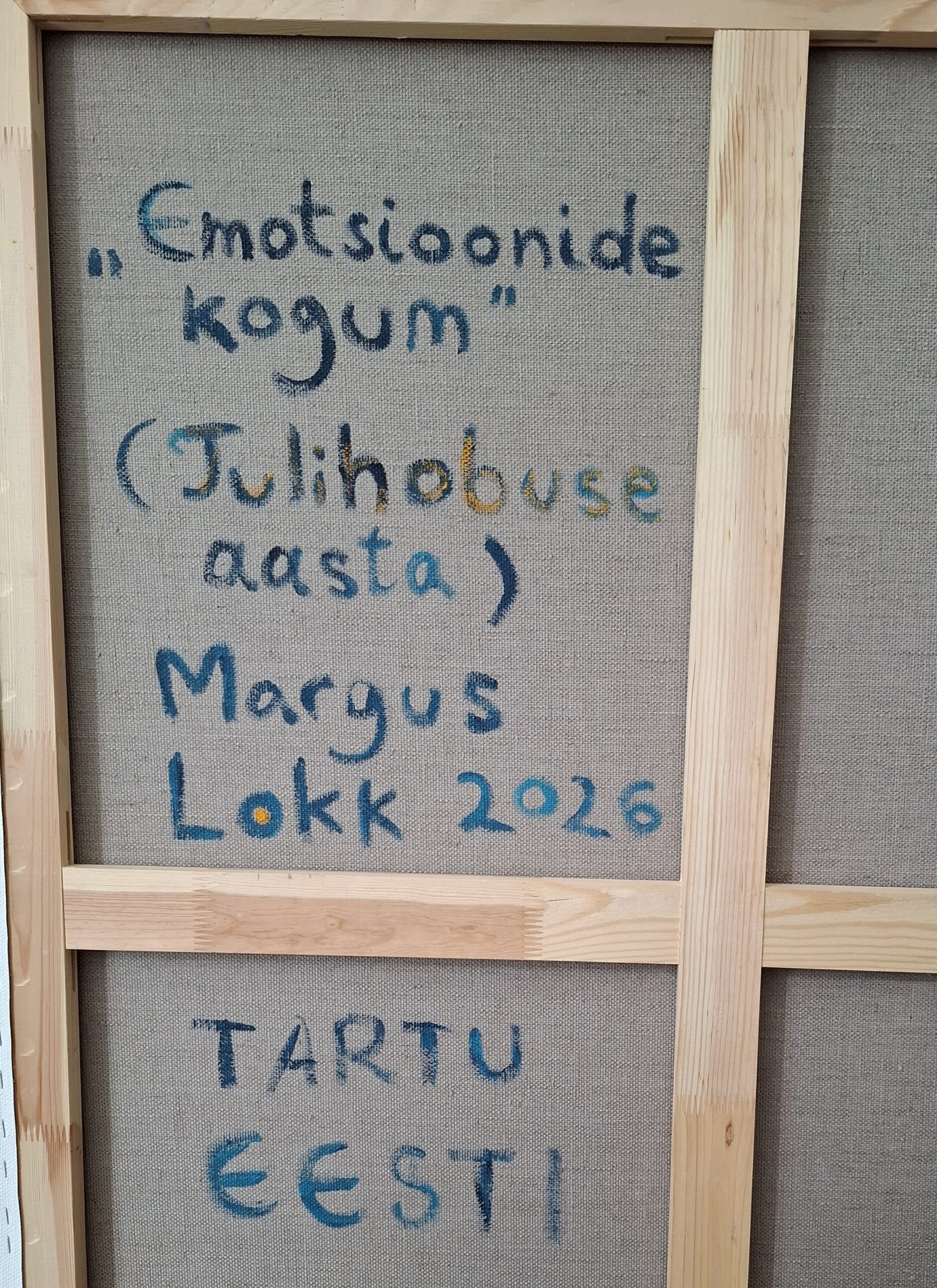 Back of a fabric art canvas with blue handwritten Estonian title 'Emotsioonide kogum' on linen, framed by wooden stretcher bars, with a signature area showing 'Margus' and date.